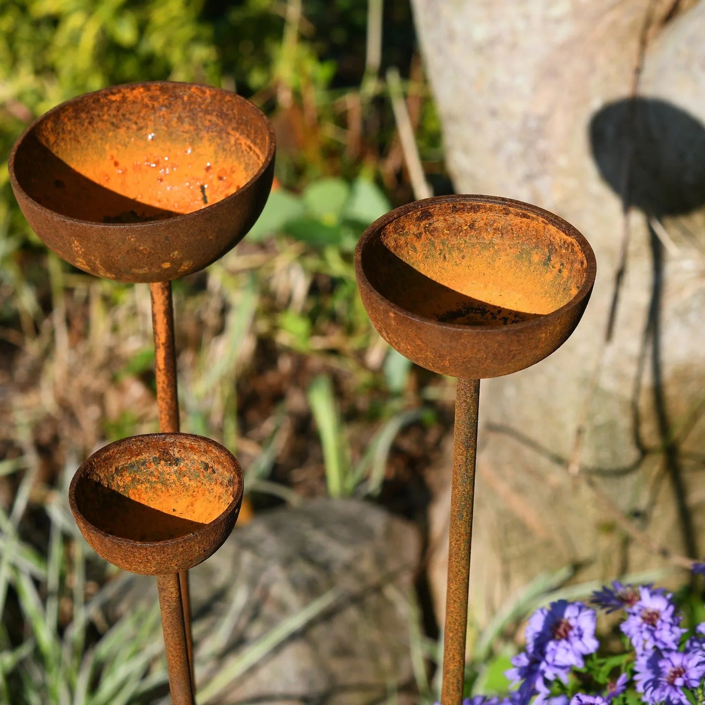 Trio of rusty metal birdbath stakes in a garden with green plants and purple flowers