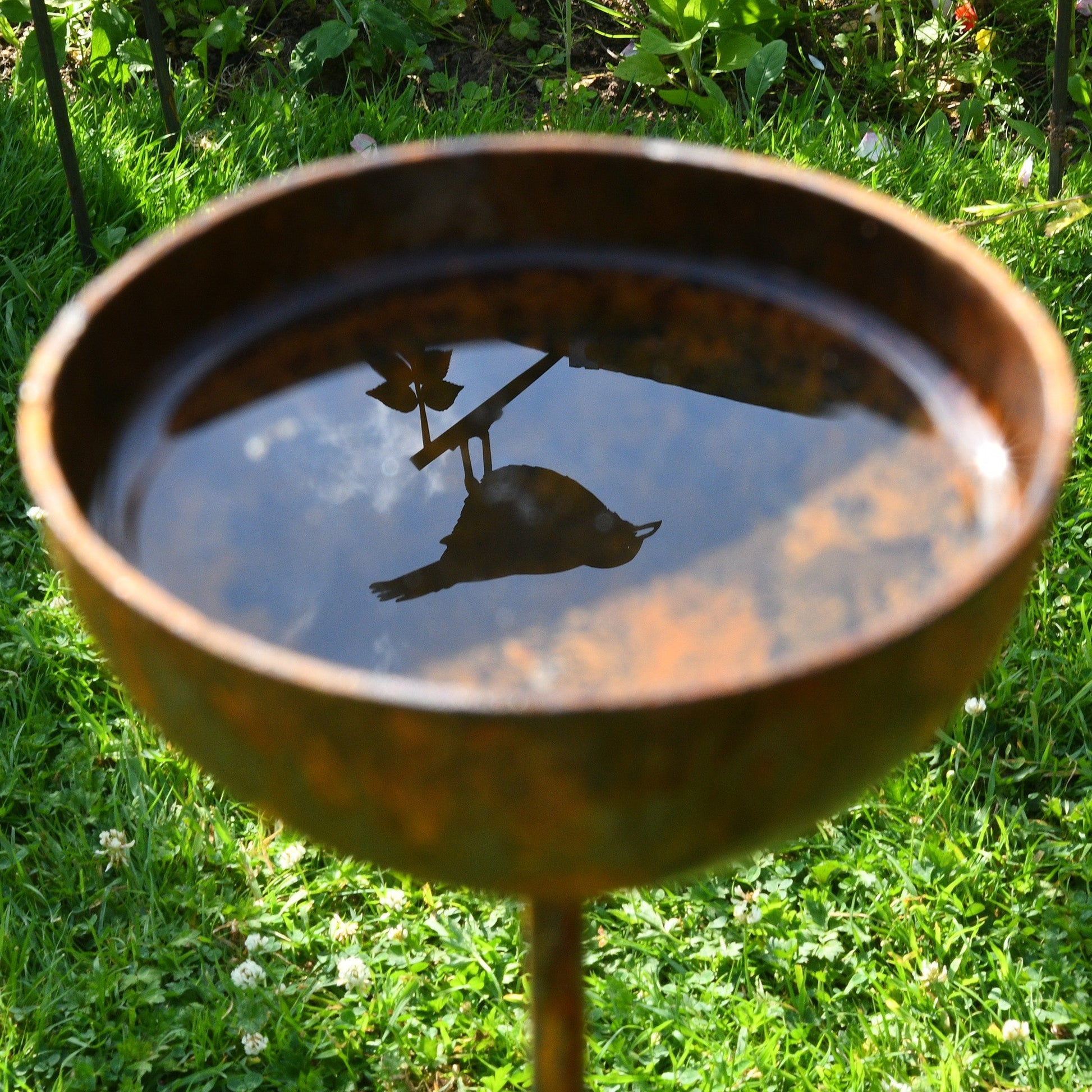 Rust-colored metal garden sculptures with shallow bowl tops, styled as birdbaths and plant supports, standing in a grassy lawn with soft floral surroundings.