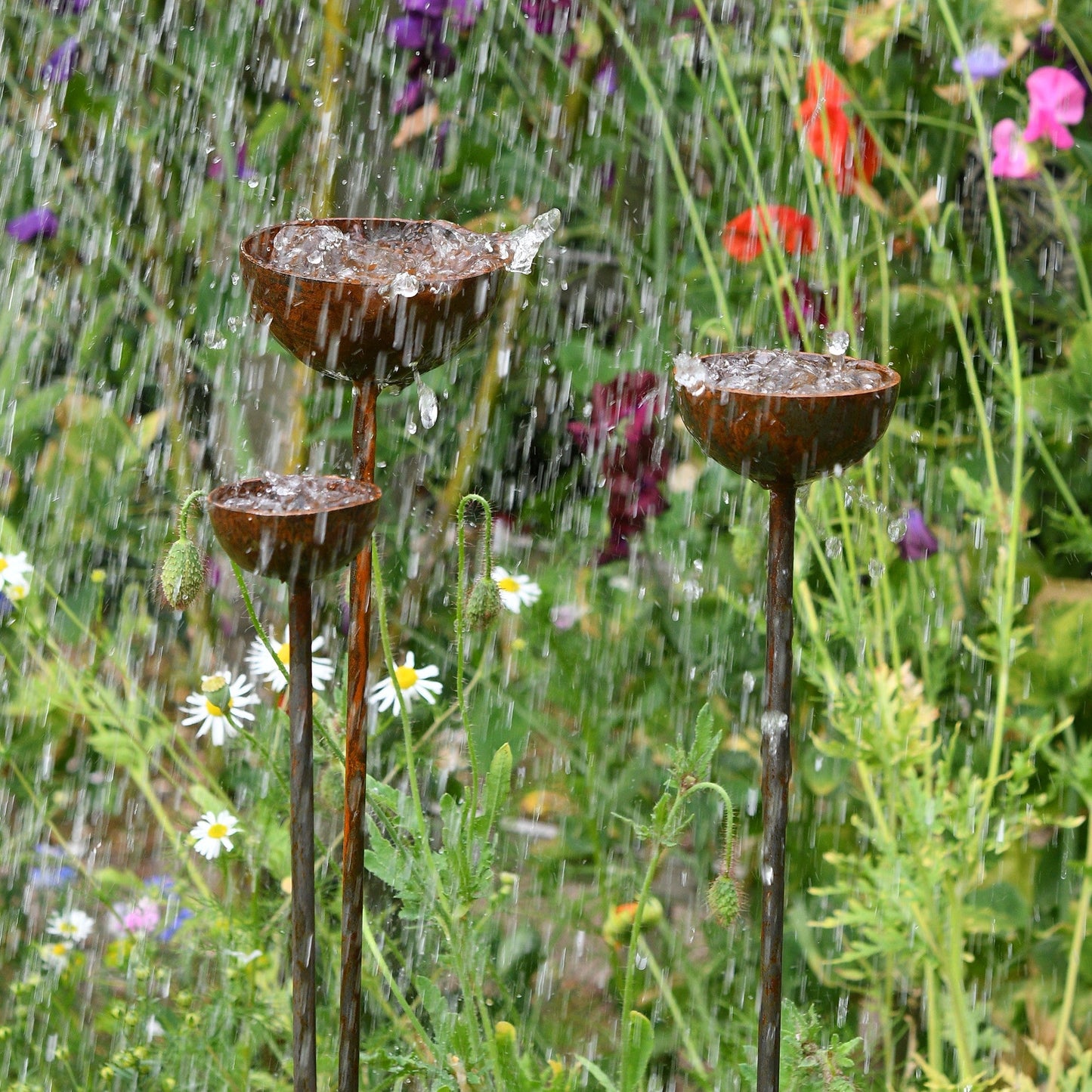 Three handcrafted rusty metal garden stakes with shallow bowl tops, styled as birdbaths and plant supports, nestled among summer flowers in a cottage garden.