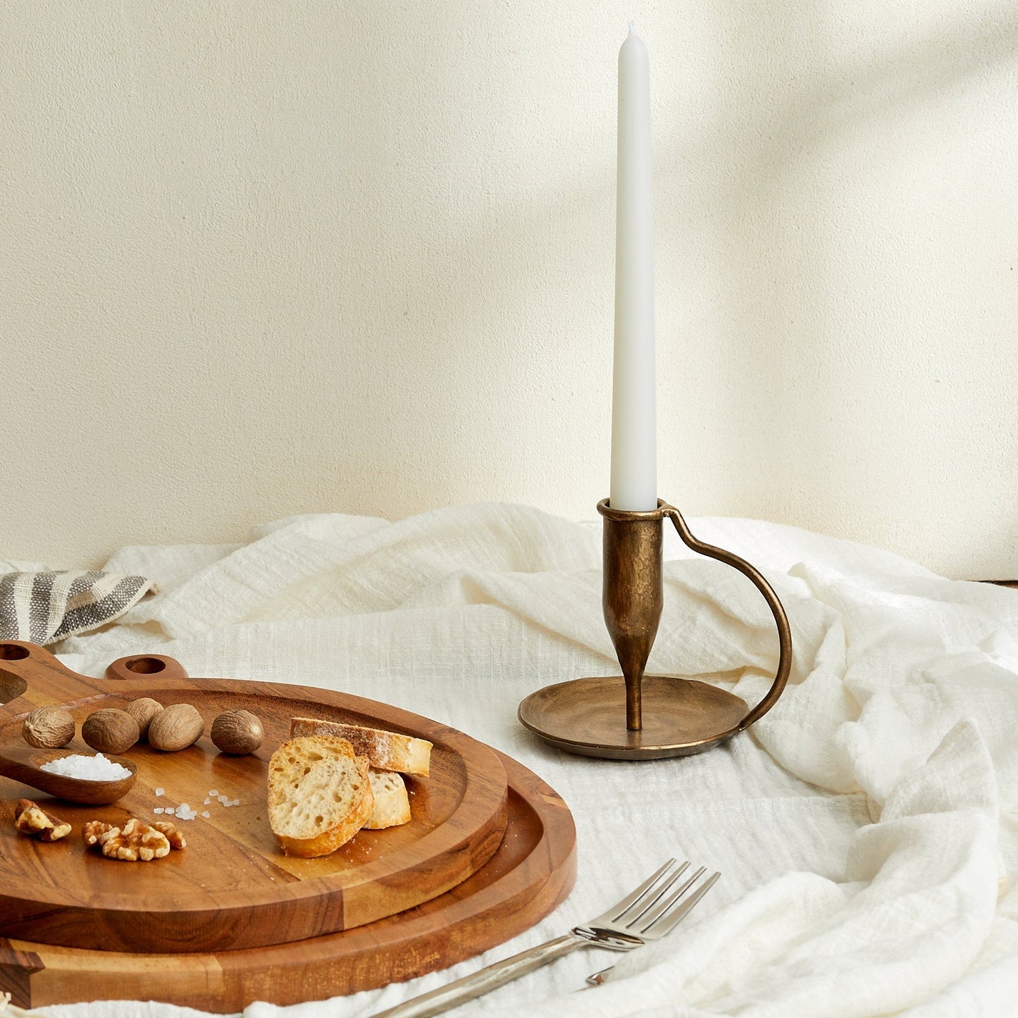 Ashbourne iron candle holder with lit taper candle on white linen tabletop, styled with rustic serving boards, fresh bread, salt pinch pots, and scattered nuts—evokes countryside warmth and timeless hosting.