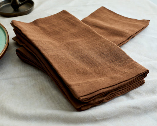 Branwell Brown muslin napkins in soft cotton, shown in a relaxed pile on a rustic table for heritage-inspired styling, soulful gatherings, and everyday rituals.