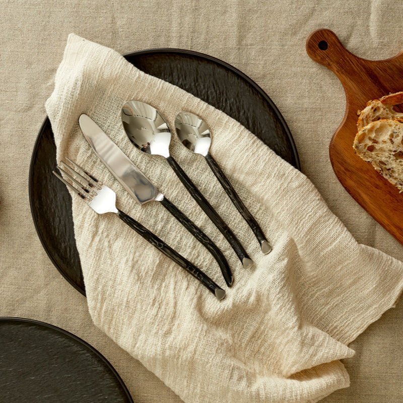Elmswell silver cutlery set styled on white linen table with black plate and folded napkin—evokes timeless elegance and soulful countryside hosting.