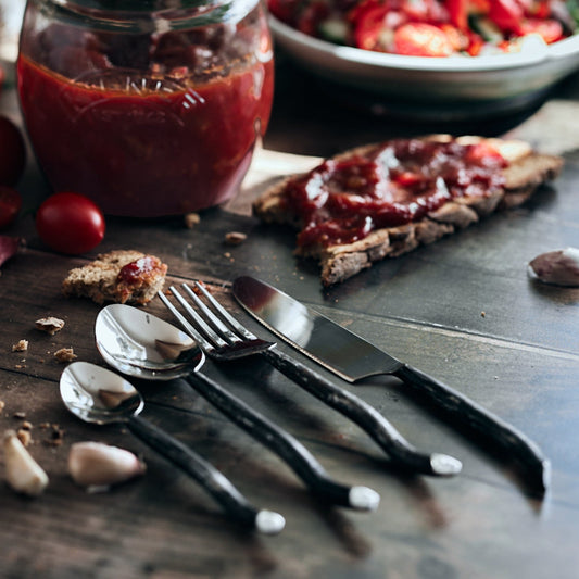 Elmswell silver stainless steel cutlery on black rustic table with sauces, bread slices, and cherry tomatoes—evokes timeless hosting and countryside charm.