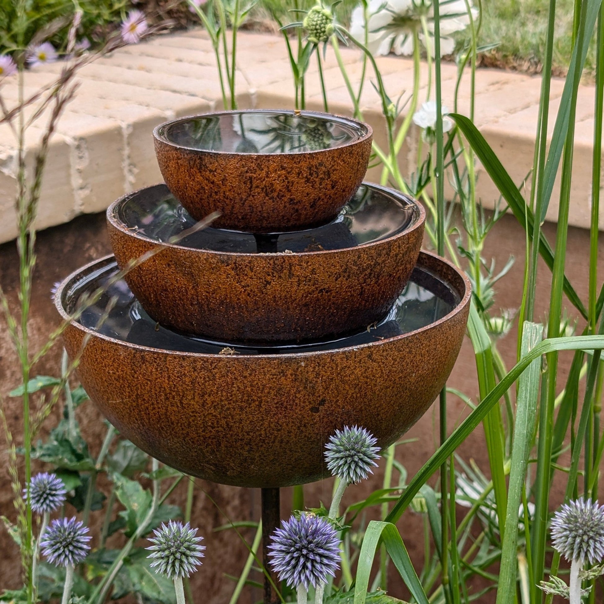 Rusty rain catcher fountain with layered metal bowls, warm orange patina, sculptural plant support and bird feeder for heritage-inspired garden styling.