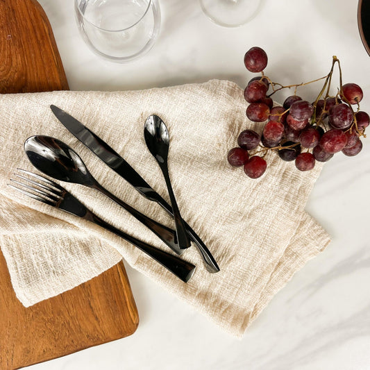 Ravenswell matte black cutlery set on marble tabletop with linen napkin, grapes, and acacia serving board—evokes rustic elegance and soulful countryside hosting.