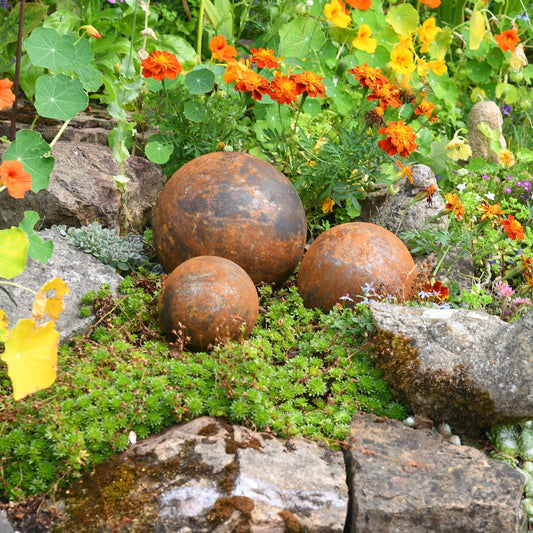 Rusted steel garden ball trio placed among flower borders, handcrafted outdoor sculptures from The Timeless Trading Co. for heritage-inspired garden styling.