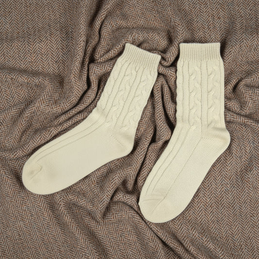Soft cream cable knit cashmere sleep socks resting on a herringbone throw in a cosy countryside-inspired setting.