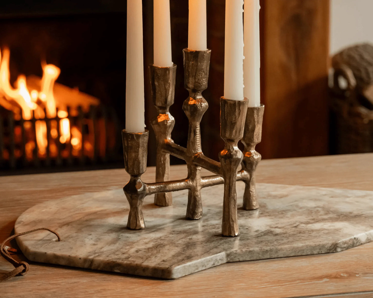 Brass candle holder with five white candles on marble board in cozy room with lit fireplace