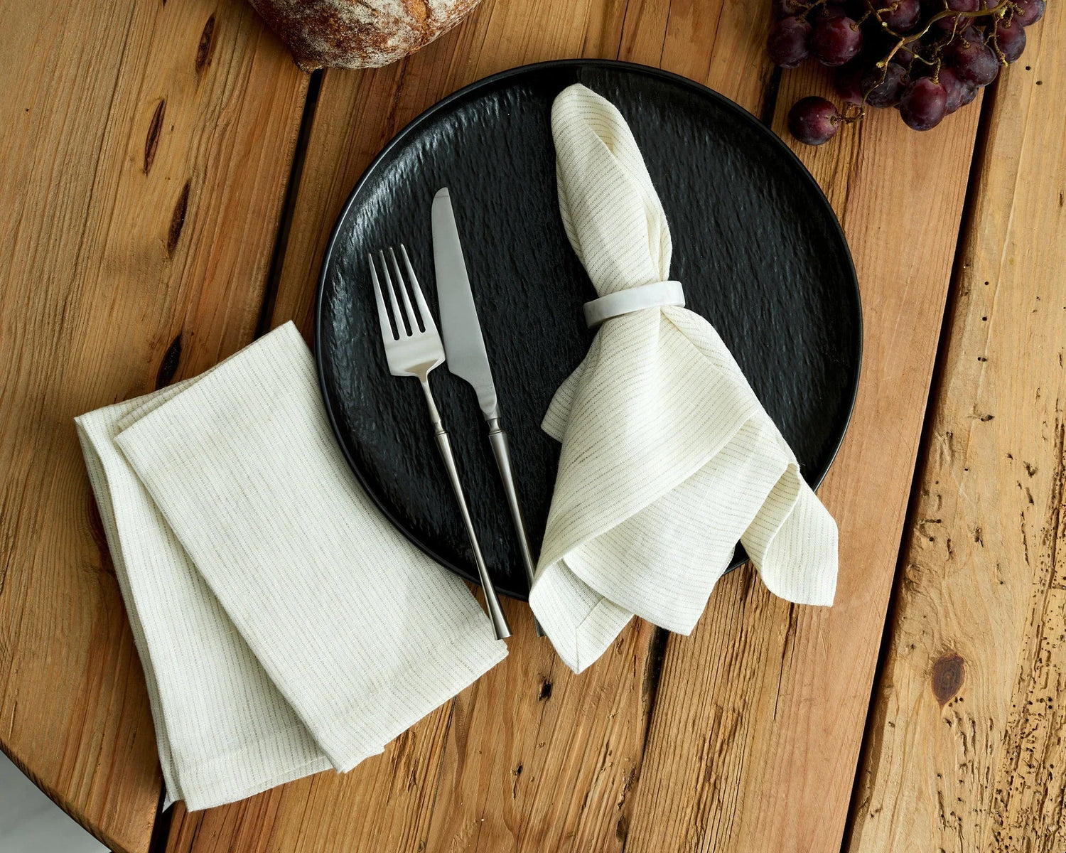 Rustic wooden table with black textured plate, white cloth napkin with ring, silver cutlery, grapes, and artisan bread