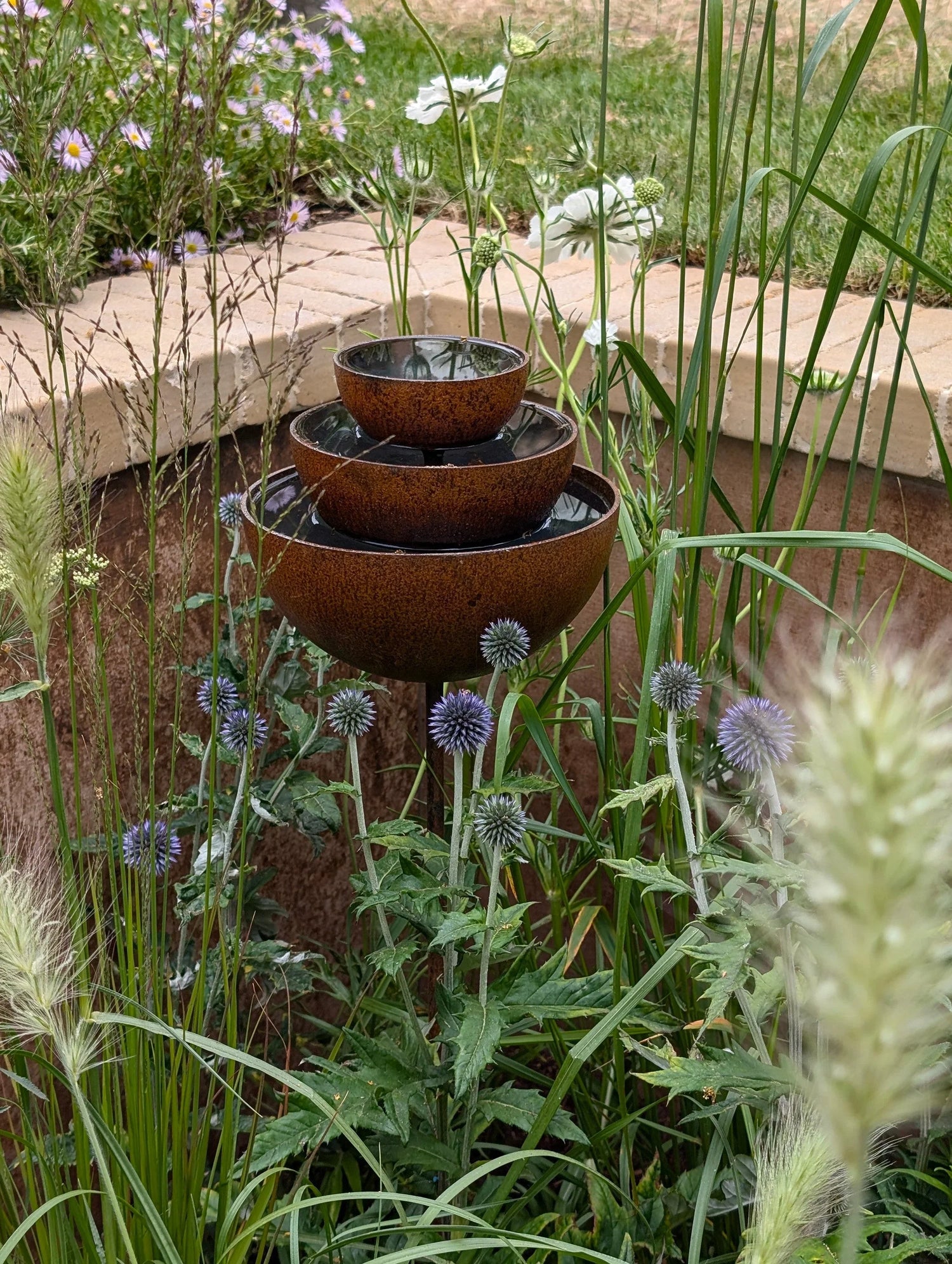 Three-tiered rustic garden water fountain surrounded by green plants and purple globe thistle flowers