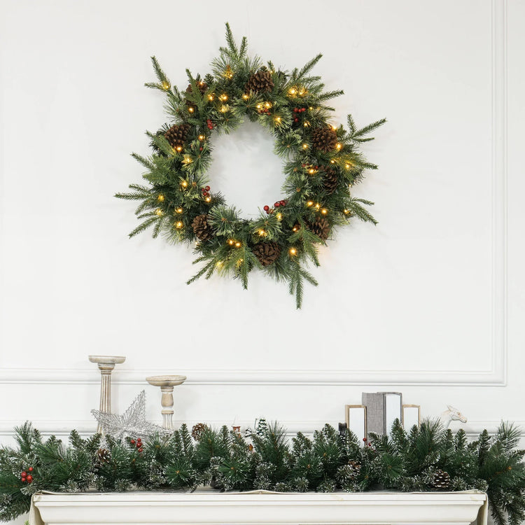 Festive green pine wreath with pinecones, red berries, and warm white lights on white wall above decorated mantel