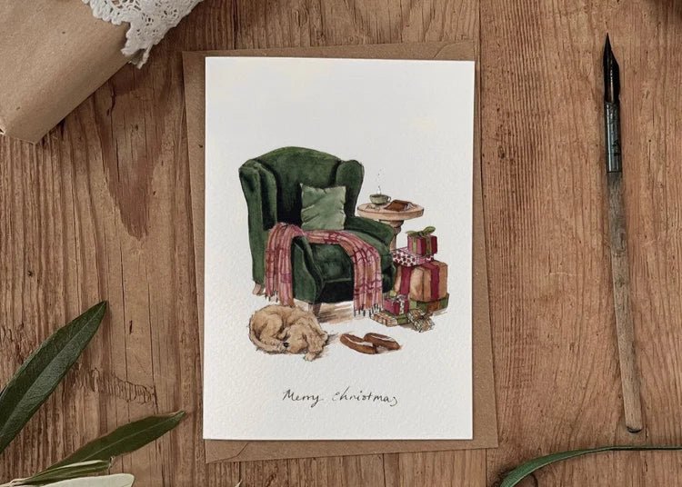 Illustrated Christmas card featuring a cosy green armchair, dog curled up on floor and wrapped gifts, — evokes countryside calm and seasonal warmth.