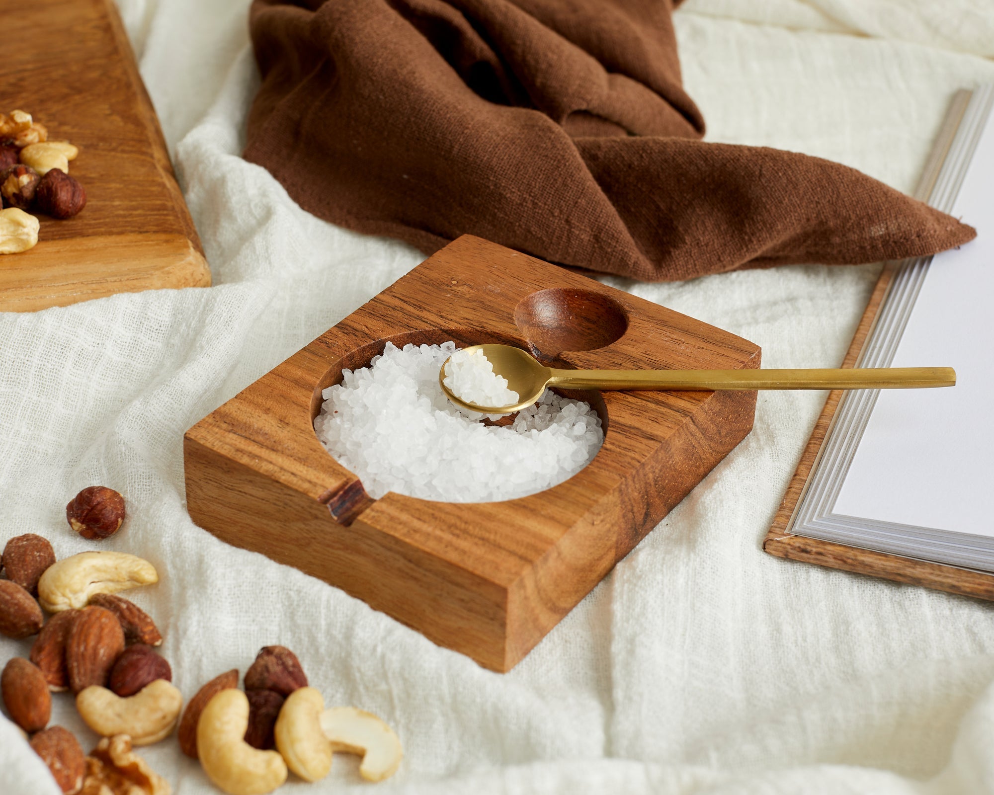 Alternate angle of acacia wood seasoning pot with brass spoon on linen with nuts and salt