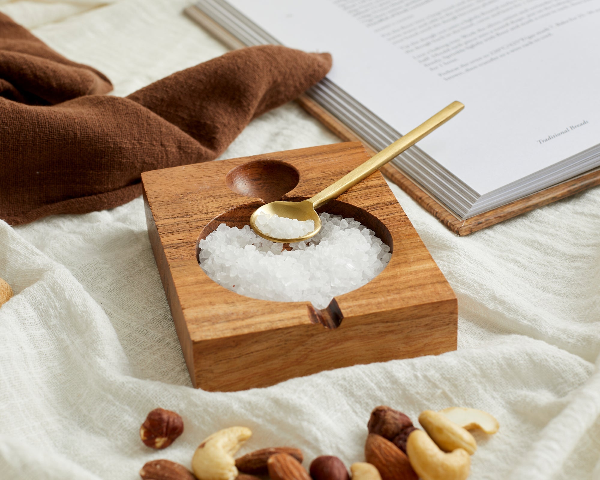 Another angle view of acacia wood seasoning pot with brass spoon on linen with nuts and salt