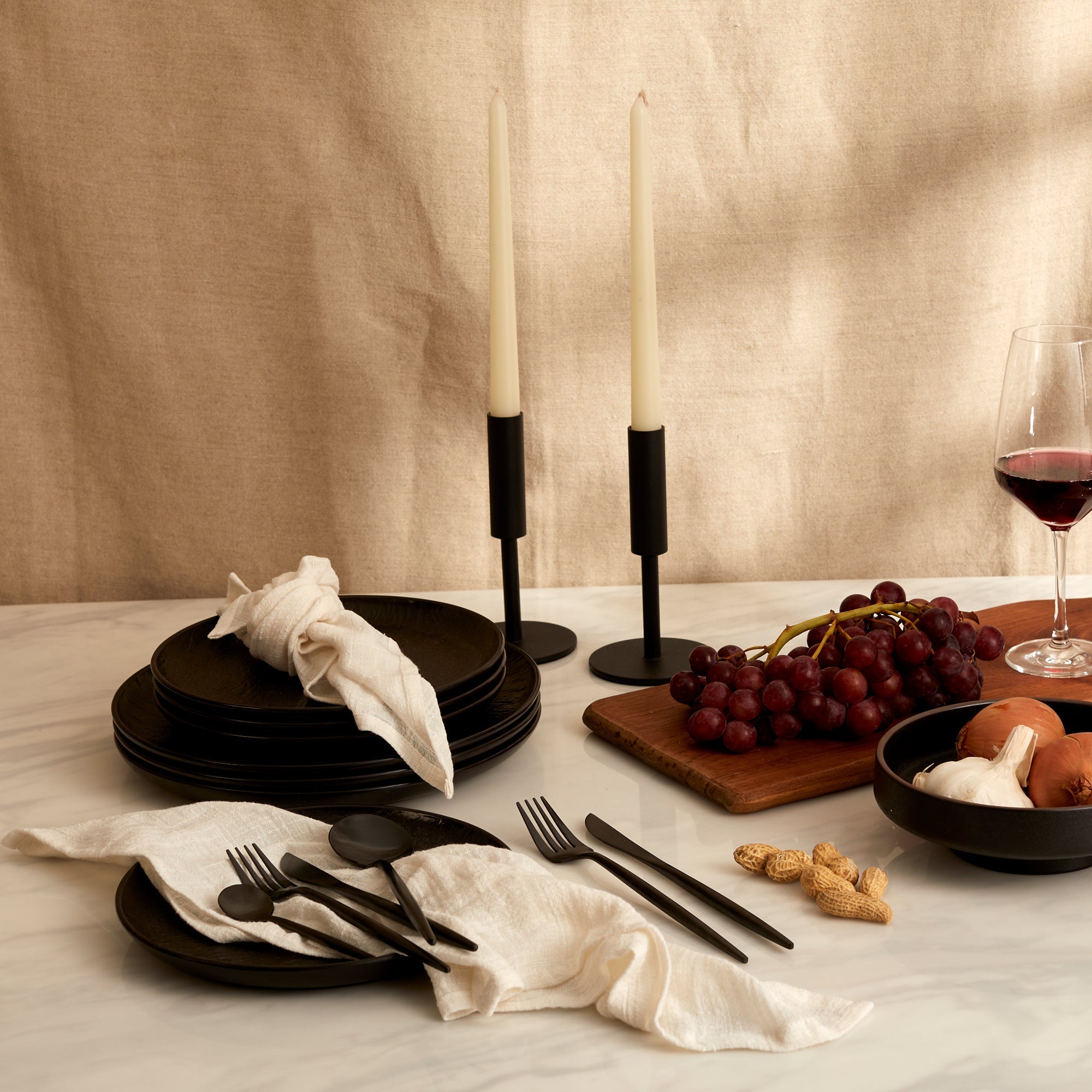 Alverley White cotton napkin set styled on a marble table with black cutlery, red wine, grapes, black candlesticks, and rustic bowls of onions, garlic, and peanuts for soulful countryside dining.