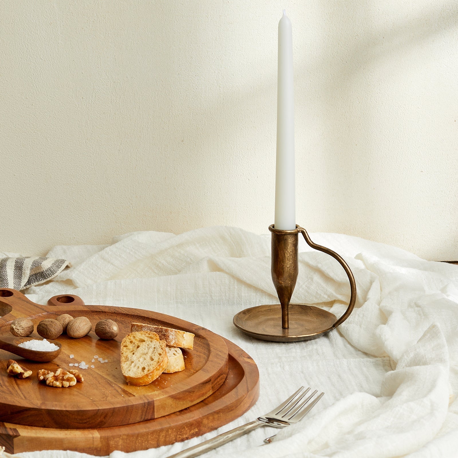 Ashbourne iron candle holder with lit taper candle on white linen tabletop, styled with rustic serving boards, fresh bread, salt pinch pots, and scattered nuts—evokes countryside warmth and timeless hosting.
