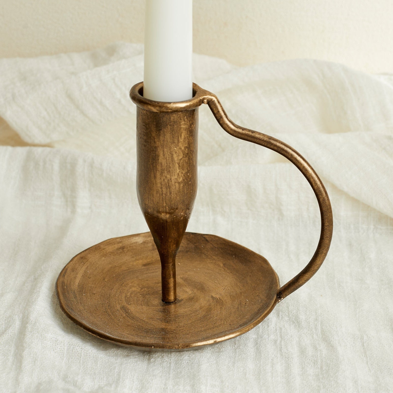 Close-up of Ashbourne iron candle holder in brushed gold finish, rustic tones glowing softly atop white linen cloth—timeless, hearth-inspired styling.