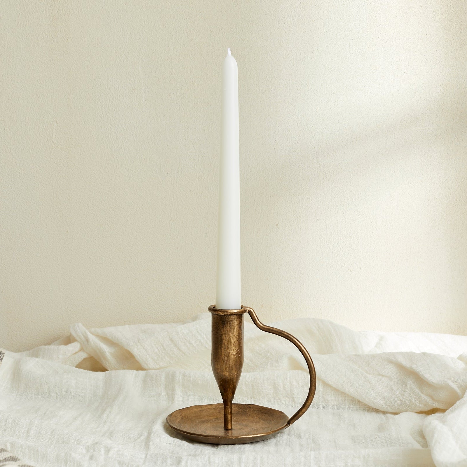 Ashbourne iron candle holder with taper candle, brushed gold tones glowing softly atop white linen—evokes rustic charm and timeless countryside ritual.