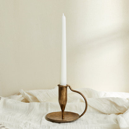 Ashbourne iron candle holder with taper candle, brushed gold tones glowing softly atop white linen—evokes rustic charm and timeless countryside ritual.